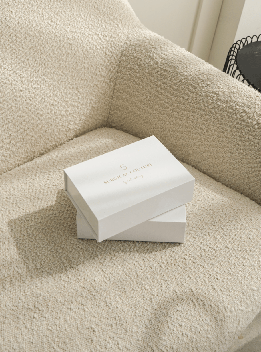 Two white ‘Surgical Couture by LaBratory’ gift boxes stacked on a textured cream armchair, bathed in natural light.