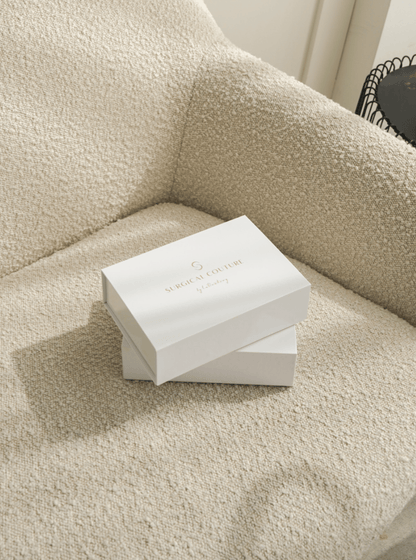 Two white ‘Surgical Couture by LaBratory’ gift boxes stacked on a textured cream armchair, bathed in natural light.